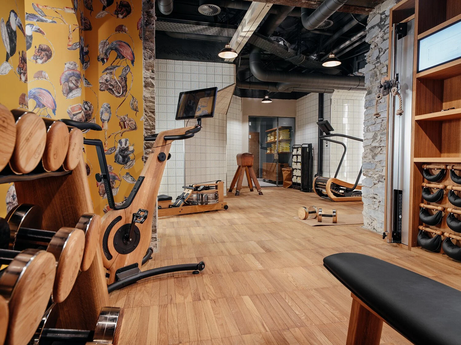 Interior of a modern gym with wooden flooring, featuring exercise equipment such as a rowing machine, stationary bike, and dumbbells. The wall on the left has patterned wallpaper with various animals. The ceiling has exposed black pipes and industrial lighting, with a stone wall on one side. There are shelves with fitness accessories and a glass door at the back.