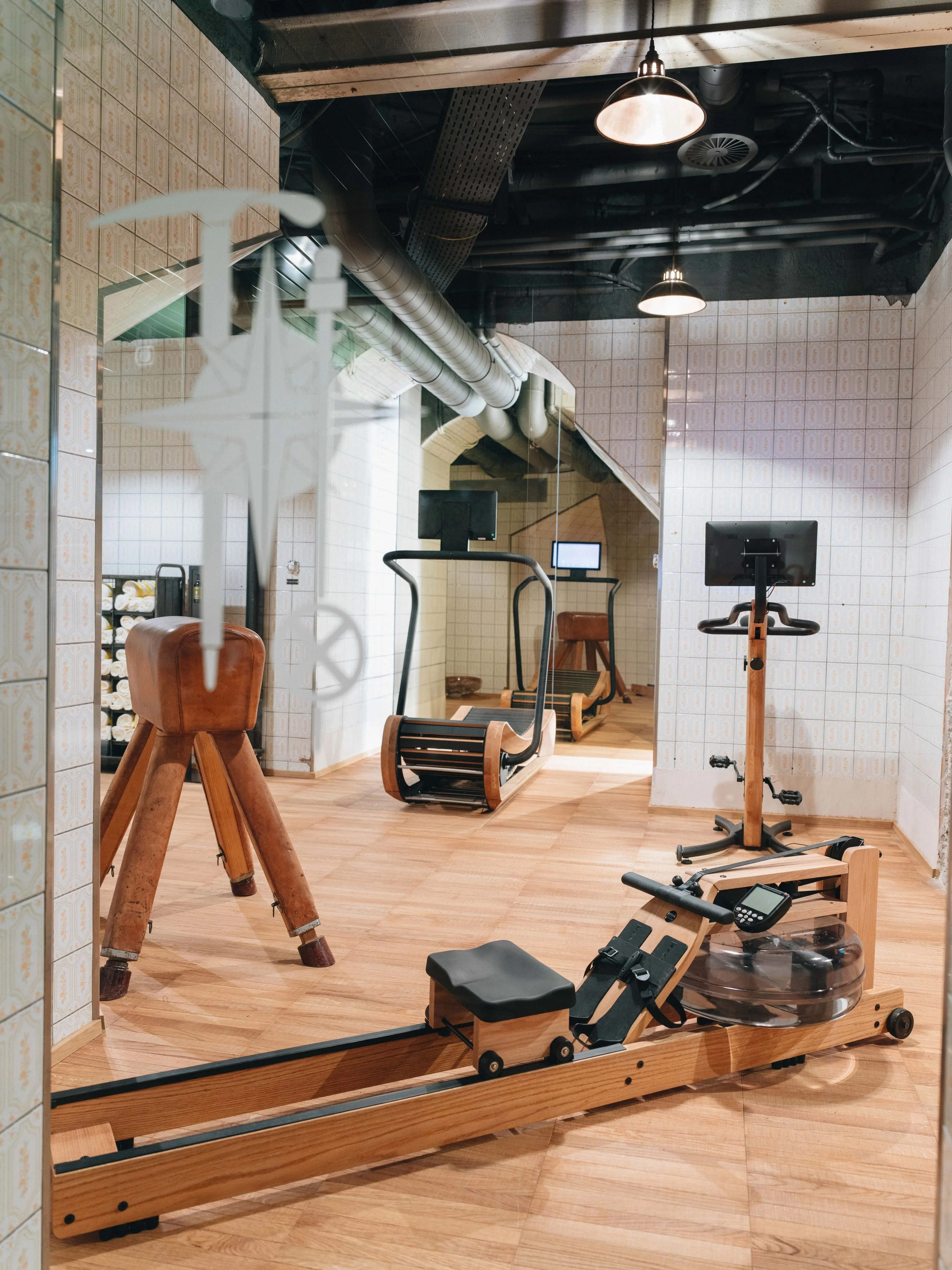This image shows a fitness or workout room with various equipment, including a wooden rowing machine, an exercise bike, a stability ball, a mirror, and some additional exercise equipment. The room has tile and wood flooring, exposed ceiling pipes, an