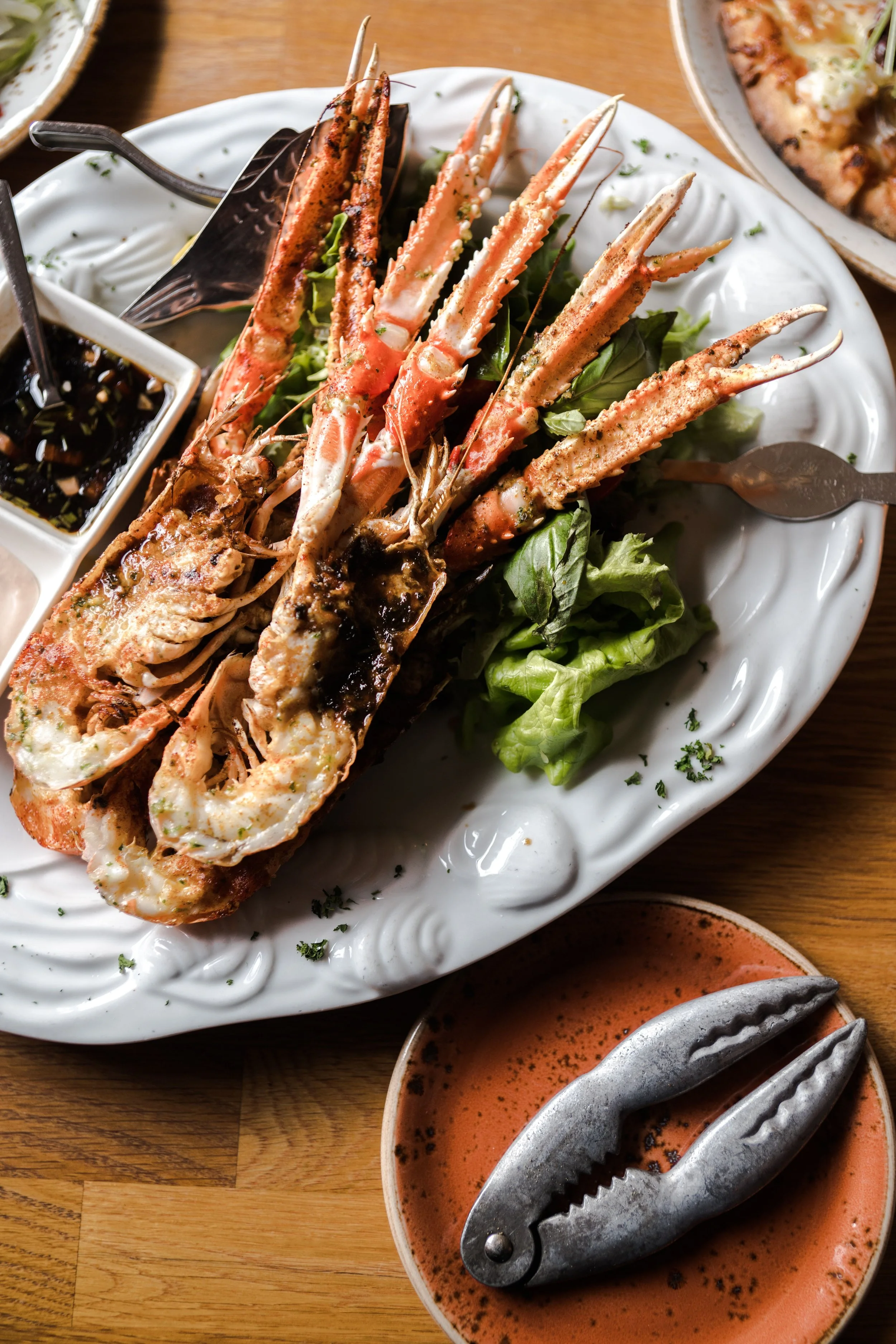 A plate of grilled lobster tails served on a bed of lettuce with dipping sauces, and a pair of seafood crackers on a nearby plate.