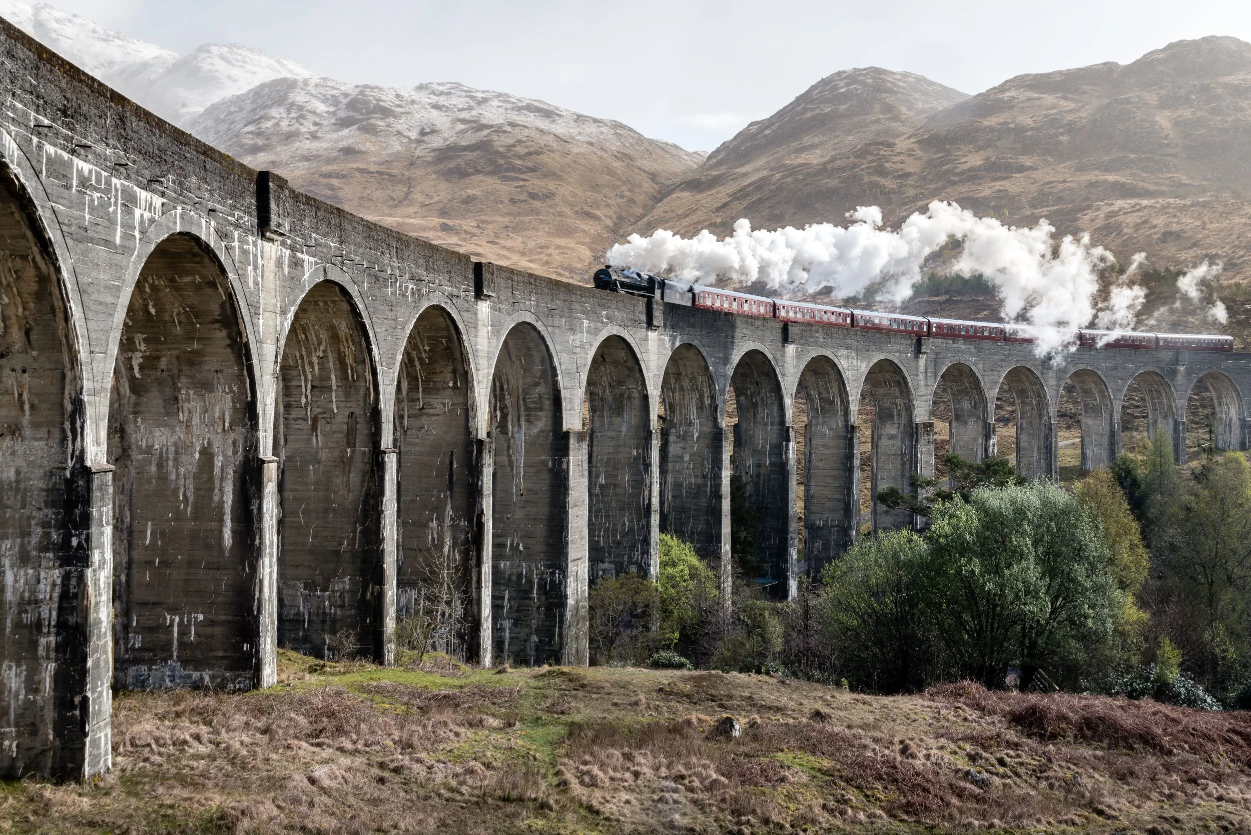 A steam train traveling across a large stone viaduct with multiple arches, set against a mountainous landscape with brown and gray hills and a cloudy sky.