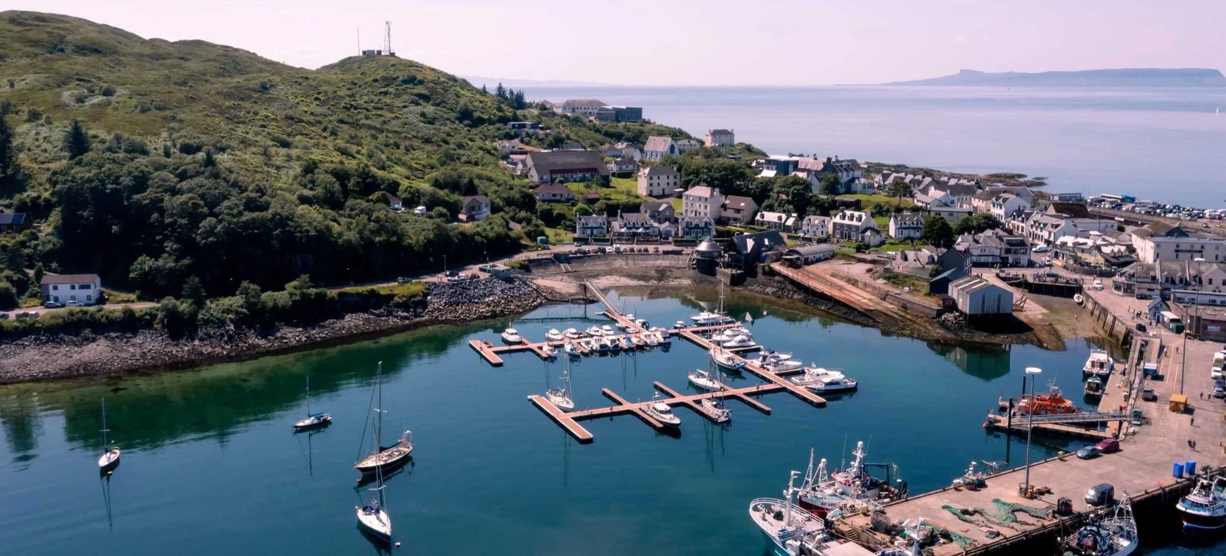 Aerial view of a small harbor with boats docked, surrounding houses on a hillside, green vegetation, and distant ocean shoreline with mountains.