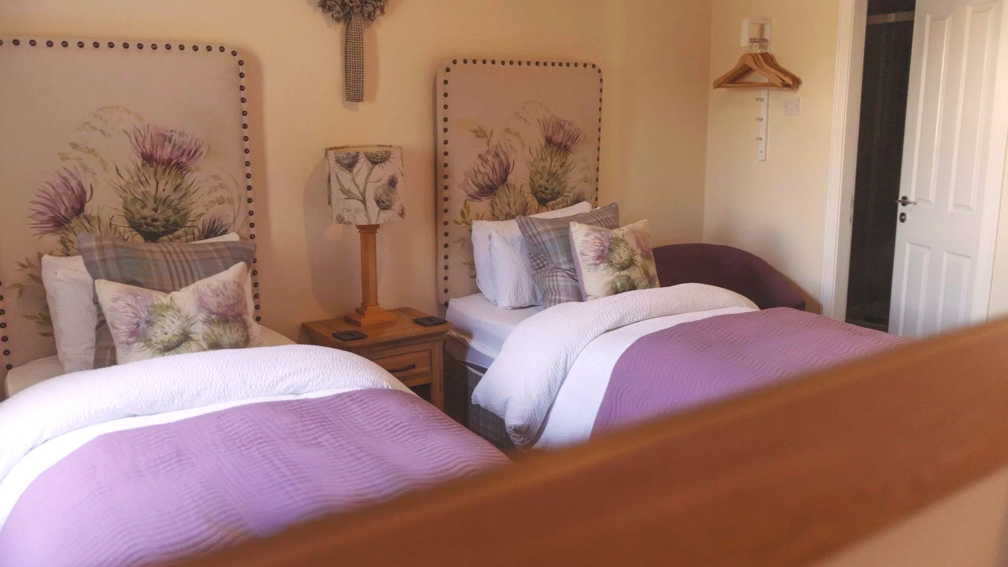 A cozy bedroom with three beds, each adorned with floral and plaid pillows, and purple bedspreads. There are decorative floral headboards, a wooden nightstand with a floral-patterned lamp, and a wall with hanging wooden hangers and a wall-mounted rail. The room has light yellow walls and a white door.