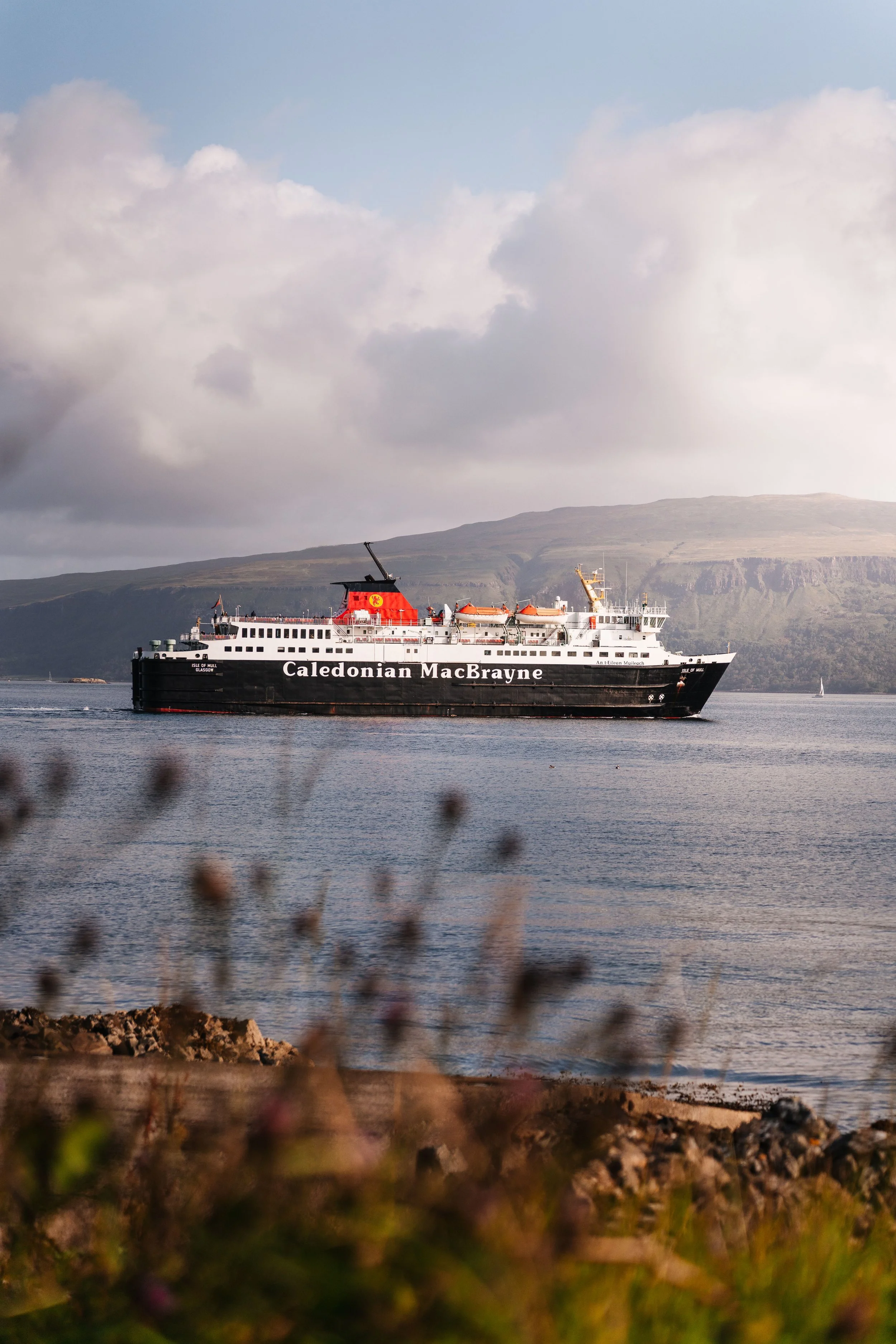 A Caledonian MacBrayne ferry boat sailing on calm water with hills in the background and a cloudy sky overhead.