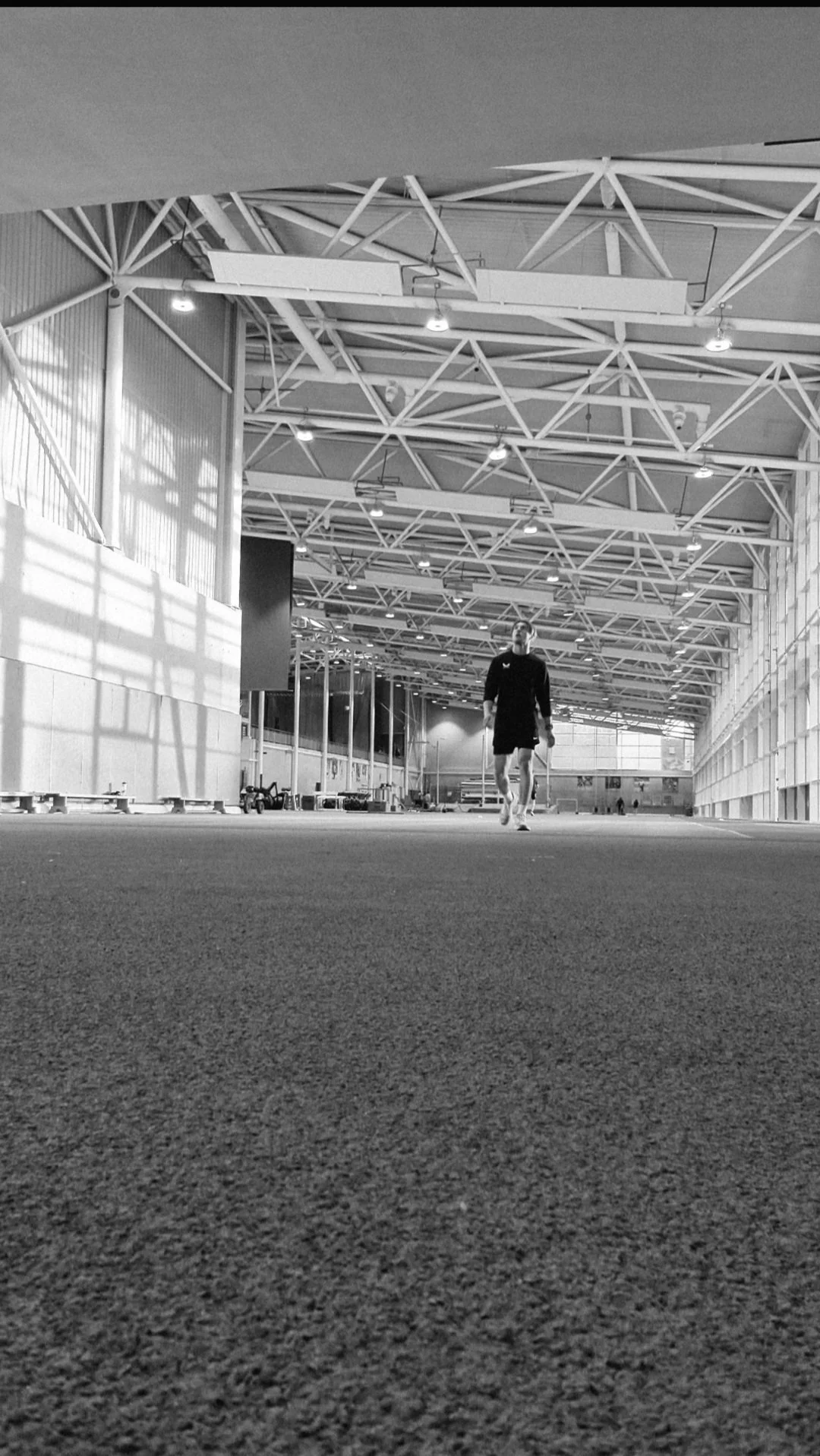 A person in athletic clothing walking inside an indoor sports facility with high ceiling, structural steel beams, and large windows, black and white photograph.