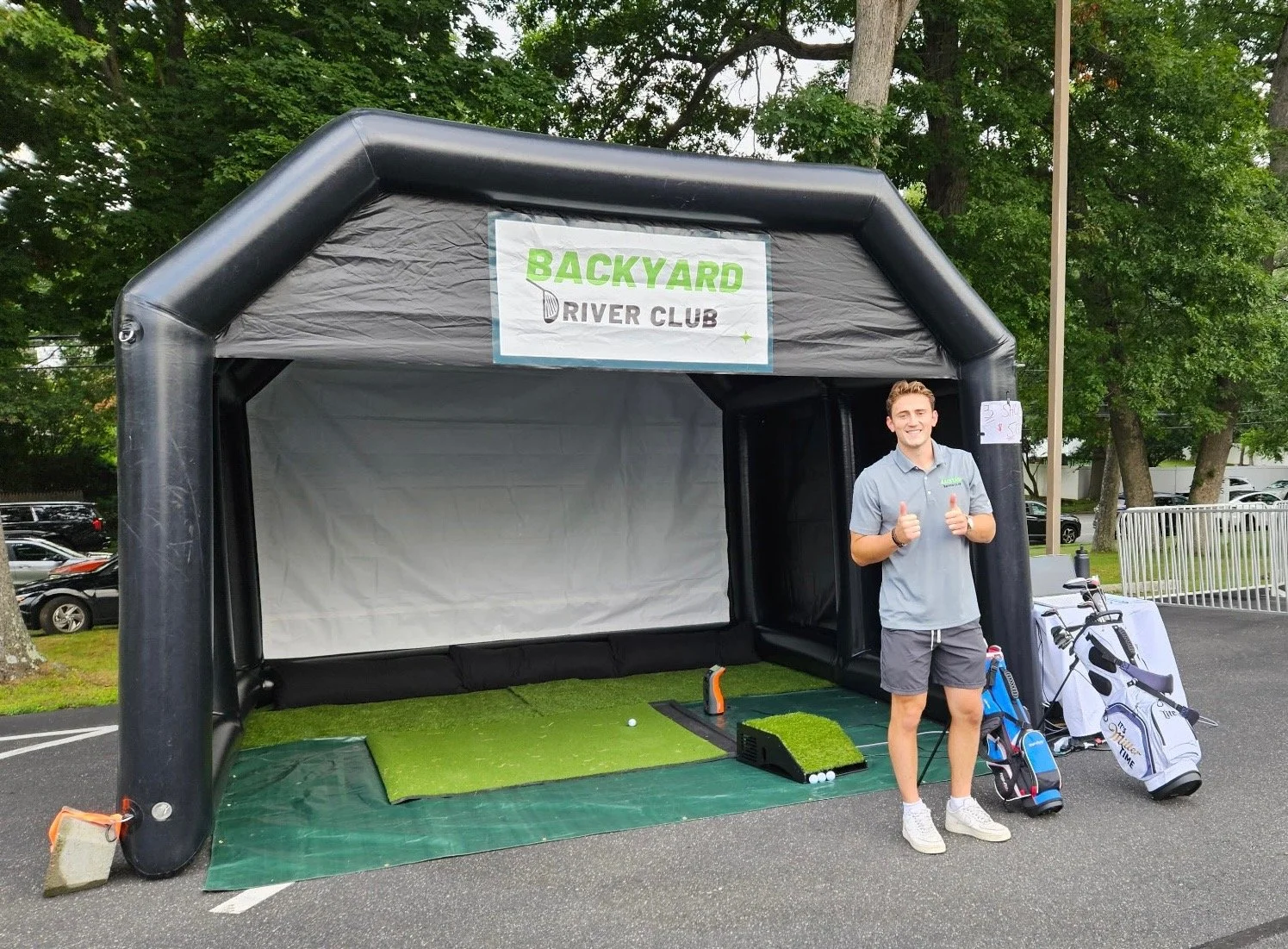 Owner of Backyard Driver Club standing in front of the mobile golf simulator. There are golf clubs and a golf bag next to him, and the setting appears to be an outdoor parking lot with trees in the background.