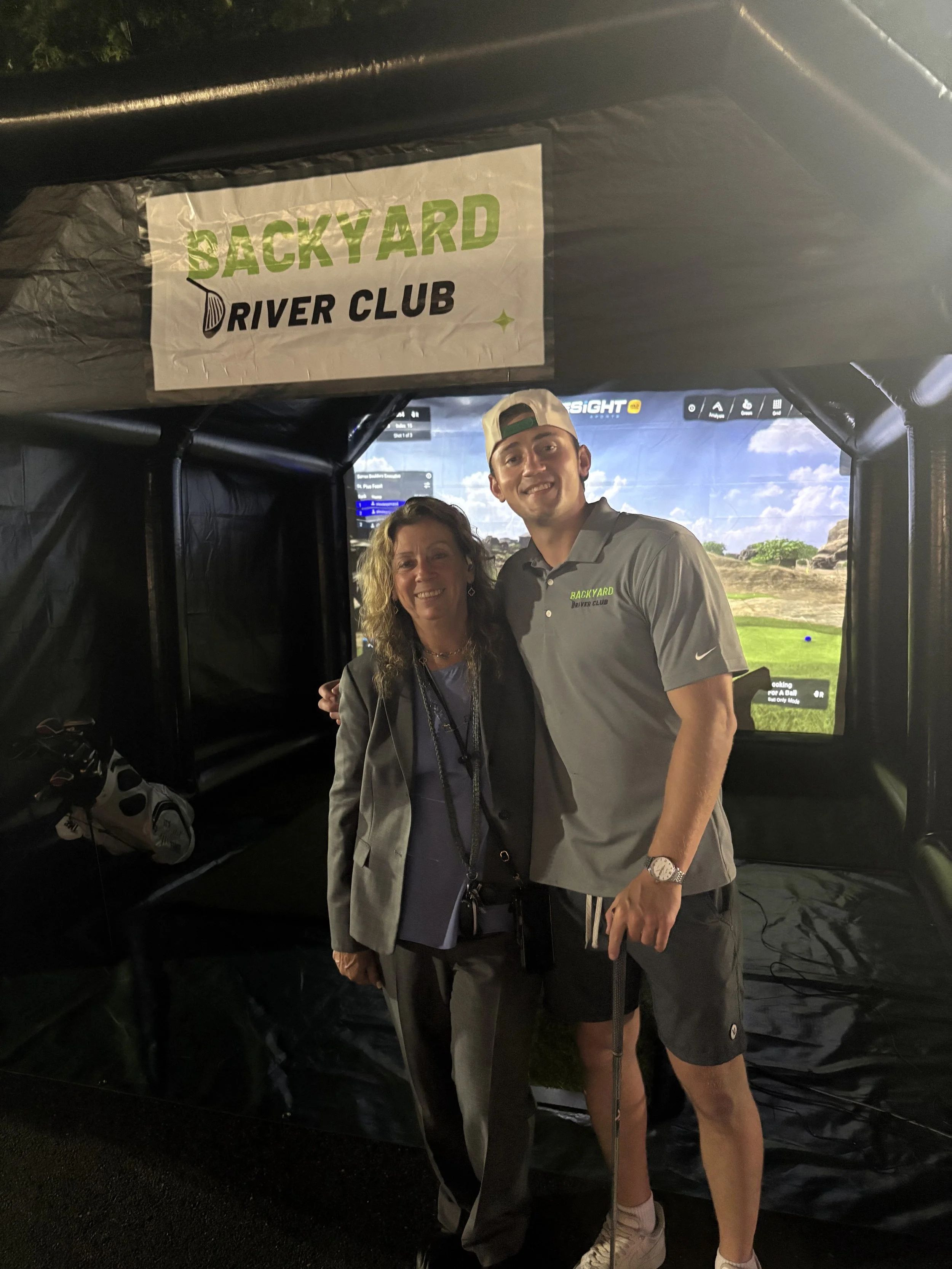 Two people standing inside a simulated golf driving range with a large screen in the background displaying a golf course, and a sign above them reading 'Backyard River Club'.
