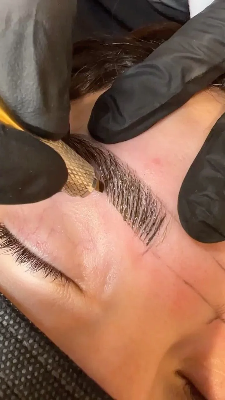 A person getting their eyebrows microbladed by a technician wearing black gloves, using a handheld tool on the person's eyebrow.