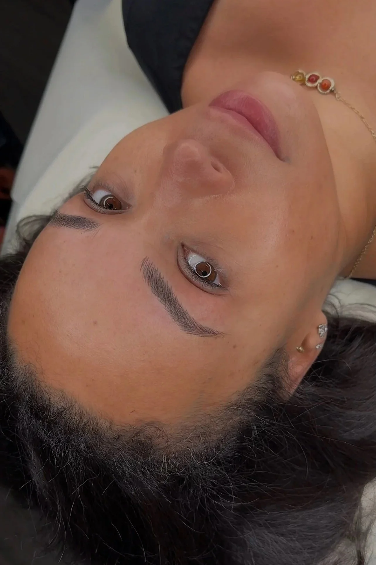 Close-up of a woman's face lying down with brown eyes, well-groomed eyebrows, and makeup, wearing a gold necklace with colorful beads and small earrings.