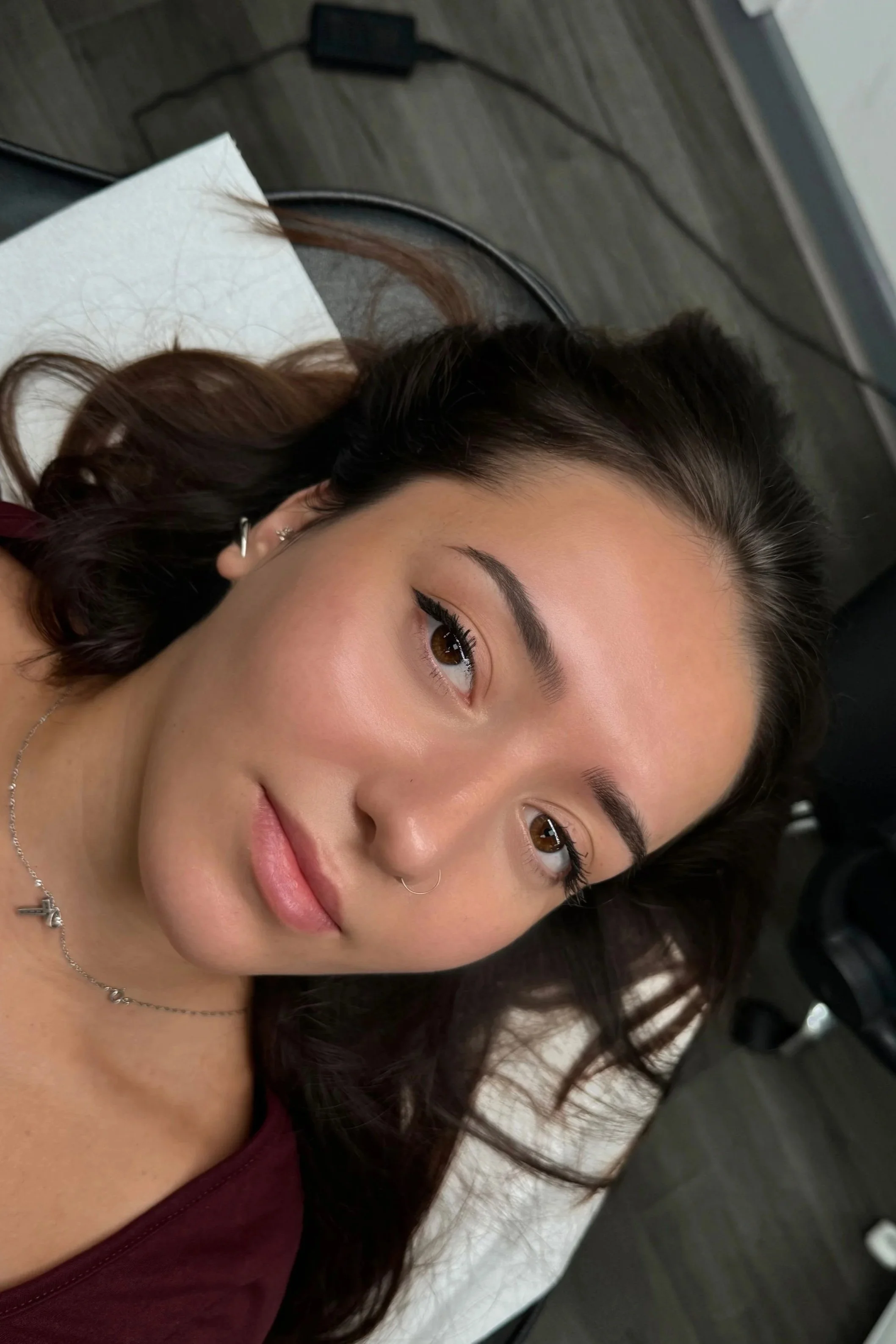A woman with dark hair and brown eyes lying on a bed, looking at the camera with a slight smile.
