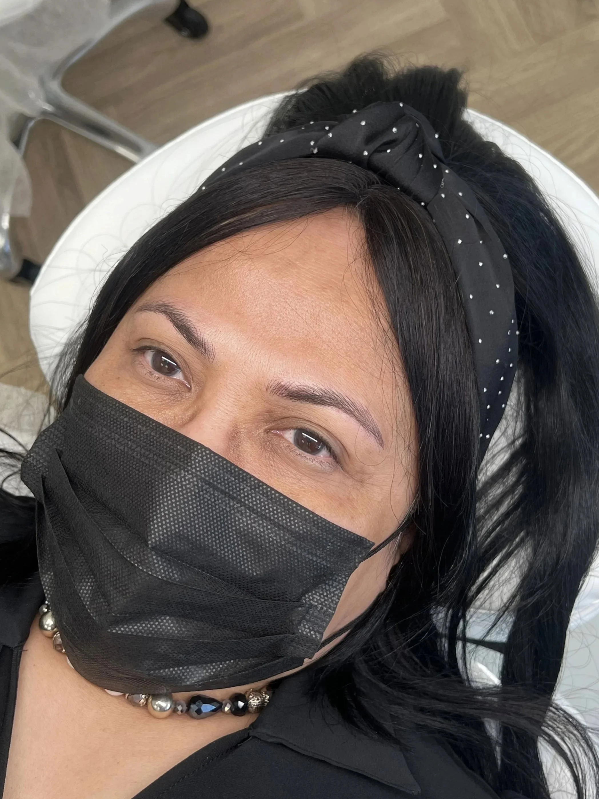 A woman with black hair wearing a black face mask, black headband with small white polka dots, and a beaded necklace, lying on a white medical chair in what appears to be a clinical setting.