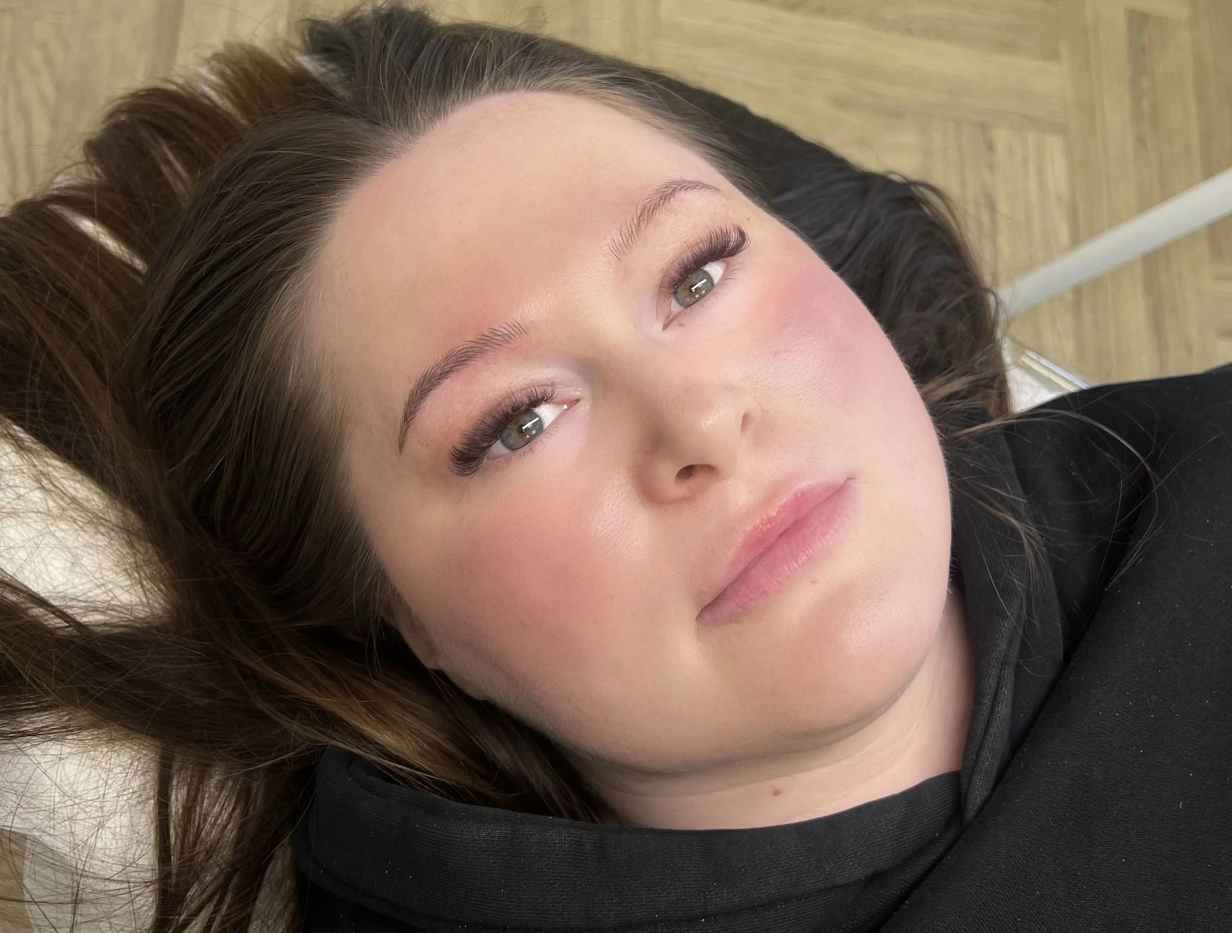 A woman lying down with her head slightly tilted and her eyes open, looking towards the camera. She has long brown hair, natural makeup, and is wearing a dark top. The background shows a wood-patterned floor.
