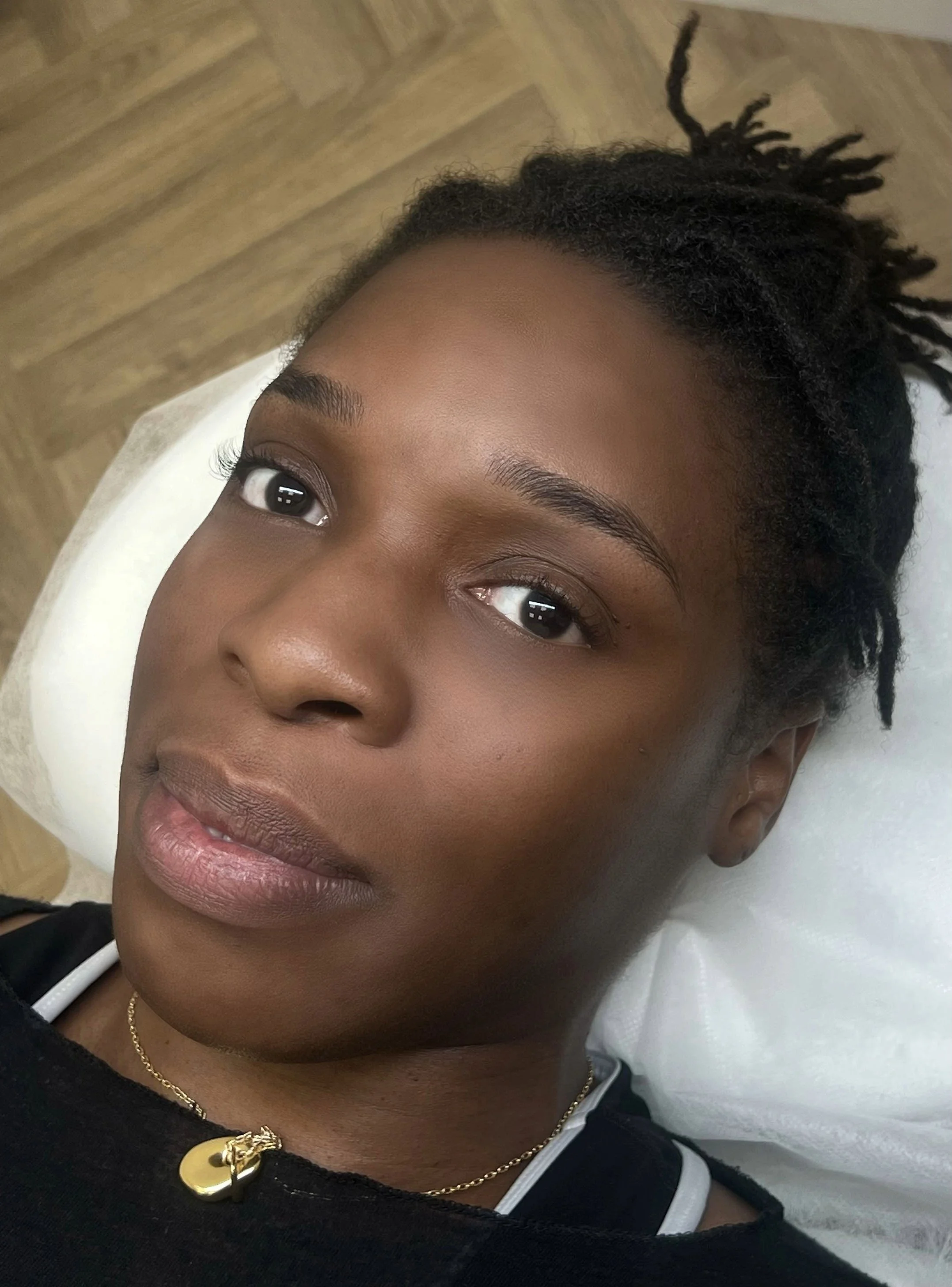 Close-up of a woman's face with smooth skin, dark eyes, and well-groomed eyebrows, lying on a white pillow with pulled-back hair styled in dreadlocks, wearing a black top and a gold necklace with a charm.