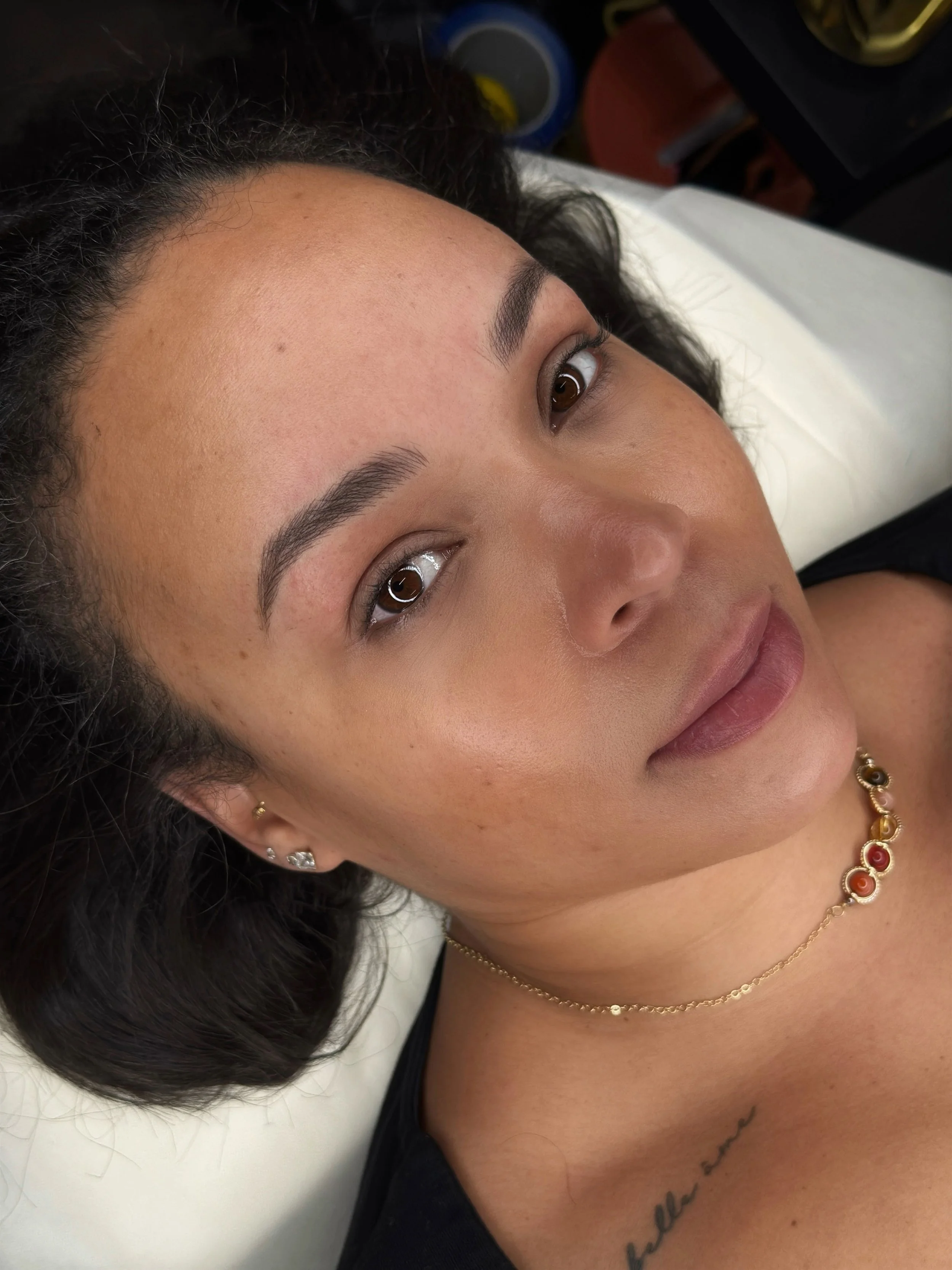 Close-up of a woman with curly dark hair lying on a white medical bed, wearing jewelry including earrings, a necklace, and a bracelet. She has a tattoo on her chest and appears relaxed.