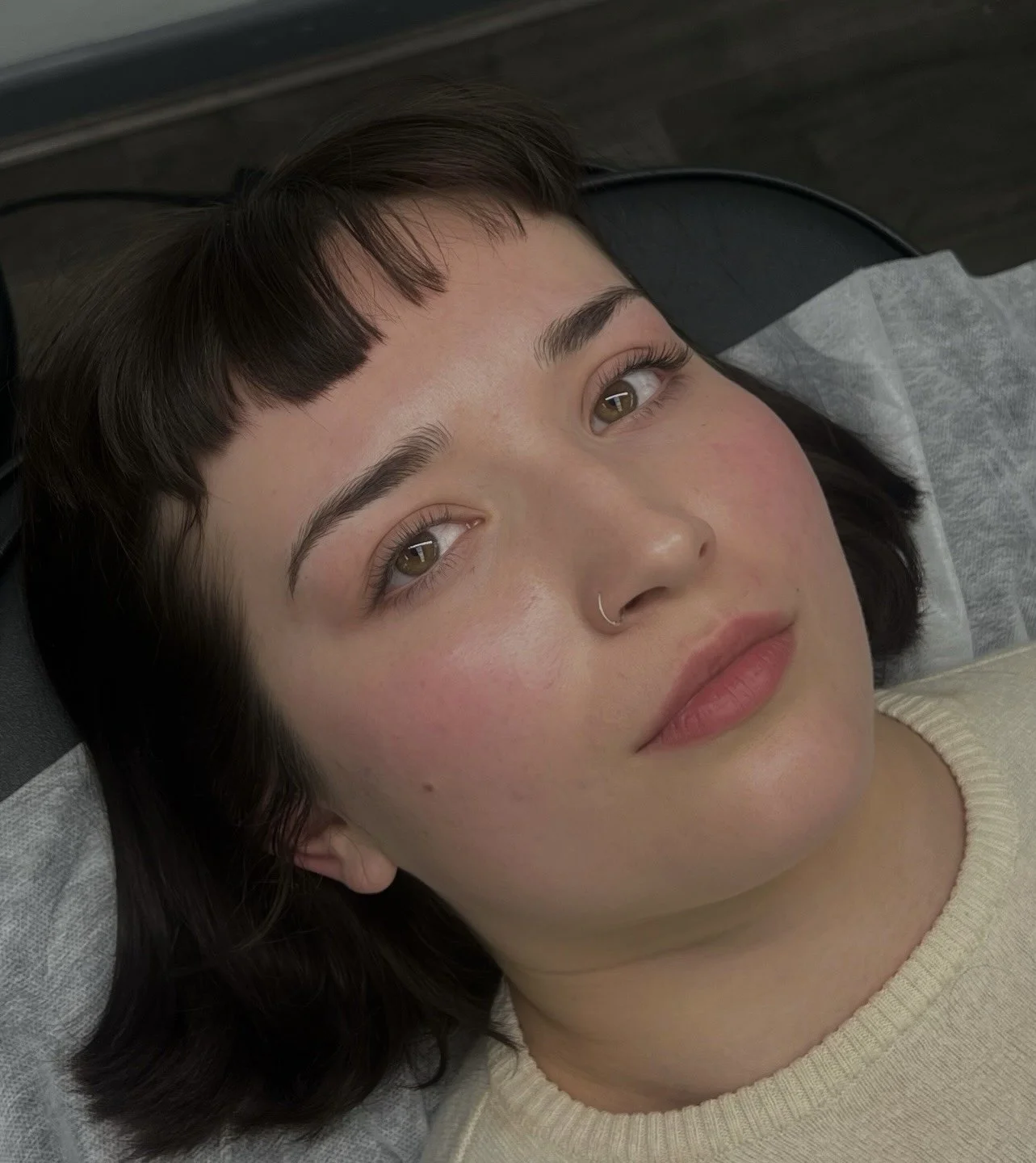 A young woman with short, dark hair and light skin, lying down with her head on a pillow, looking up at the camera. She has a nose ring and wears a light-colored top.