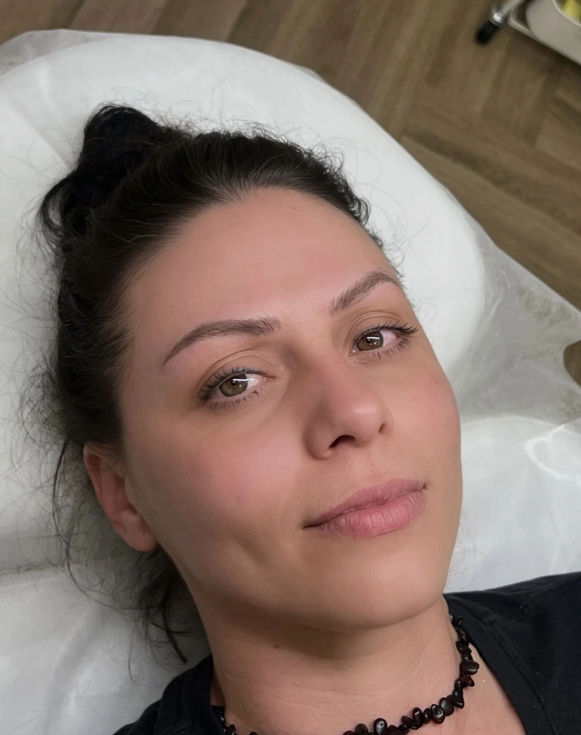 Close-up of a woman lying on a white pillow, looking up at the camera. She has dark hair styled in a bun, light skin, and is wearing a black top and a black bead necklace. The background shows a wooden floor and part of a medical or massage table.