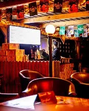 Interior of a dimly lit bar with a television on the wall, wooden decor, chairs, and bottles displayed above the bar