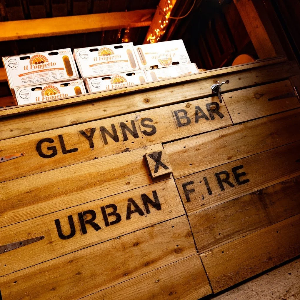 Wooden bar with the sign 'Glynn's Bar' and 'Urban Fire' painted on it, with pizza boxes labeled 'il Faghetto' on top.