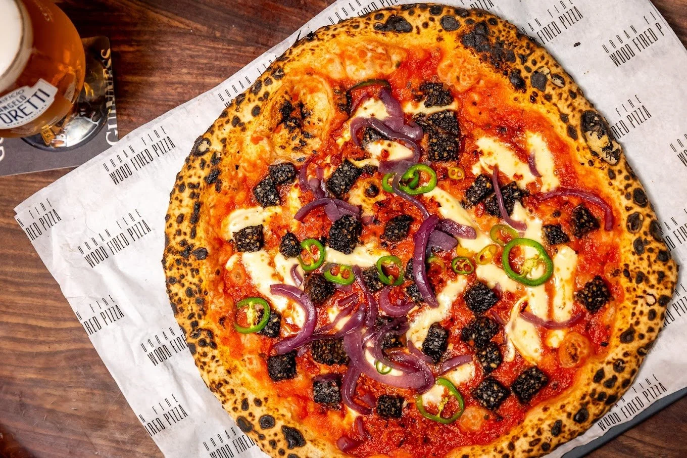 A pizza with a charred crust topped with blackened sausage, red onion, sliced green chili peppers, and melted cheese, served on branded paper on a wooden table, with a glass of beer nearby.