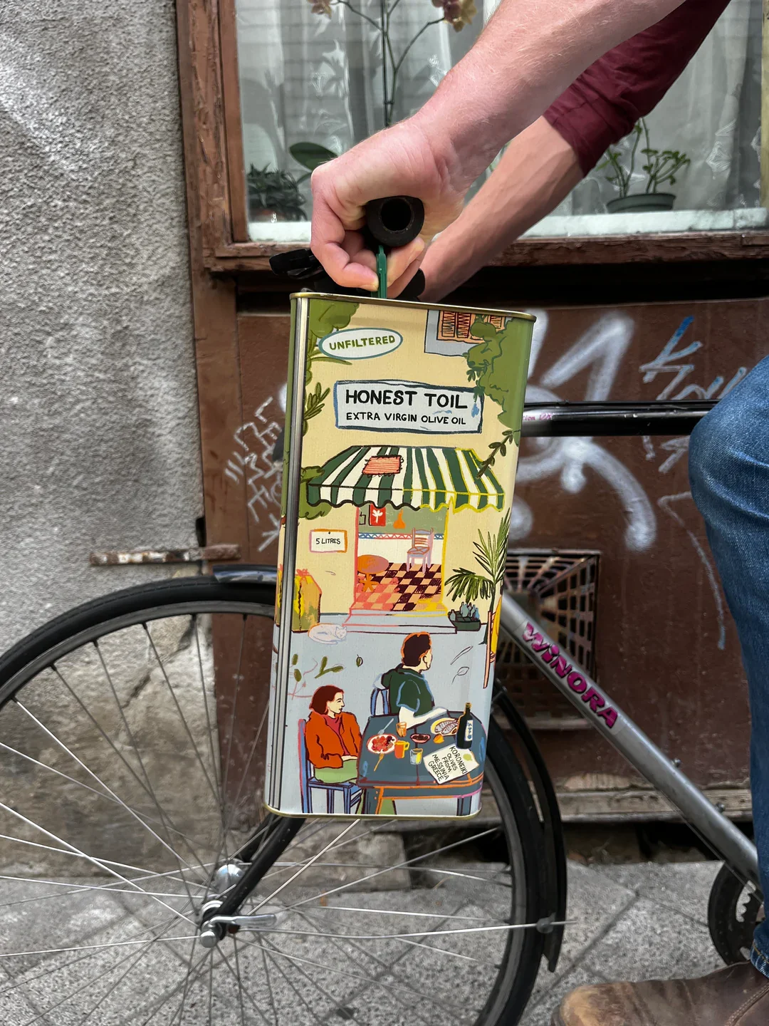 A person holding a large, colorful can of Honest Toil extra virgin olive oil with an illustrated label depicting a café scene with people sitting at a table outdoors, a bicycle wheel in the background, and a window with potted plants.