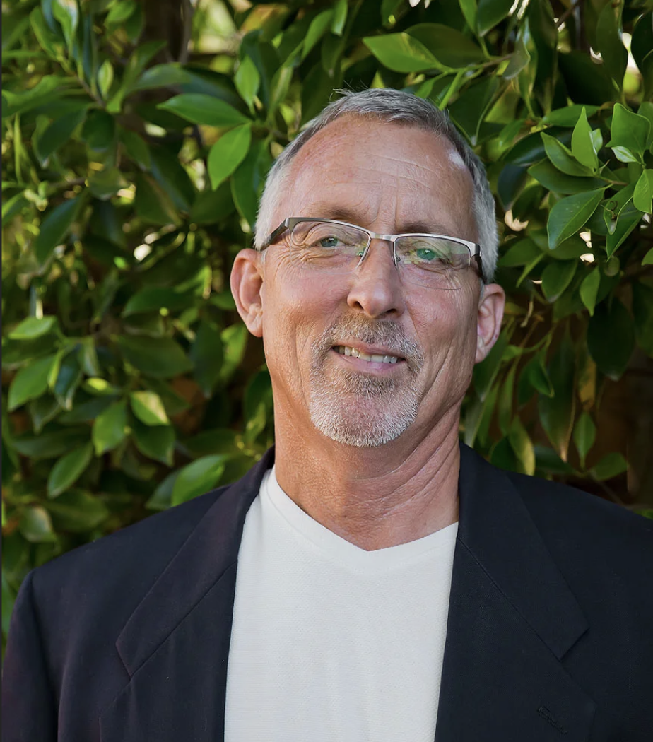 A middle-aged man with glasses and a goatee smiling outdoors, standing in front of green foliage.