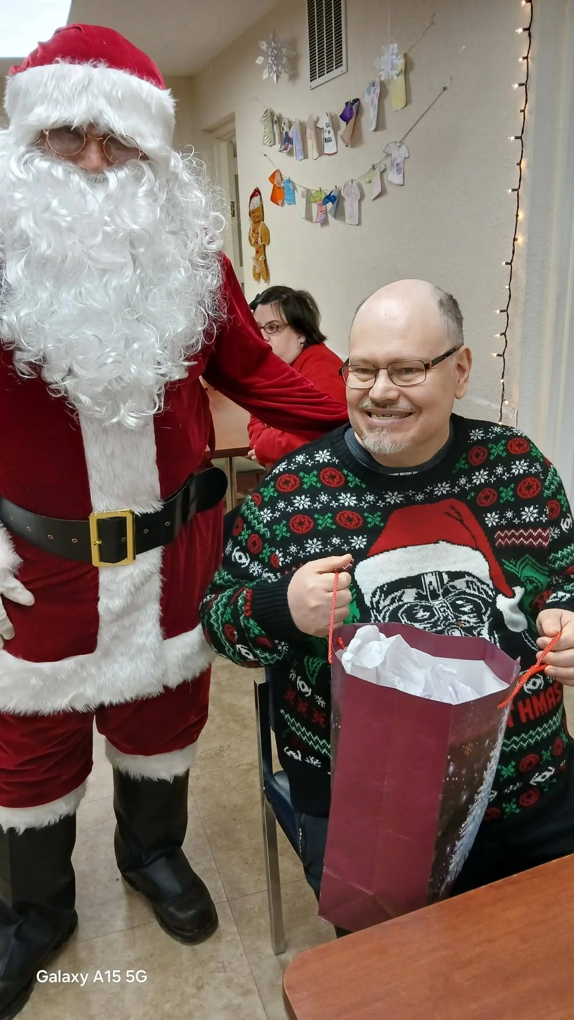 A man in a Christmas sweater sitting at a table with a gift bag, smiling, while a person dressed as Santa Claus stands next to him, inside a decorated room.