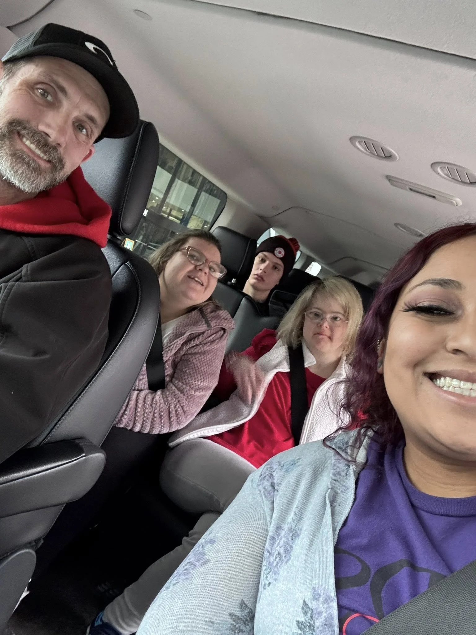 Group of five people sitting in the back of a vehicle taking a selfie, smiling and looking at the camera.