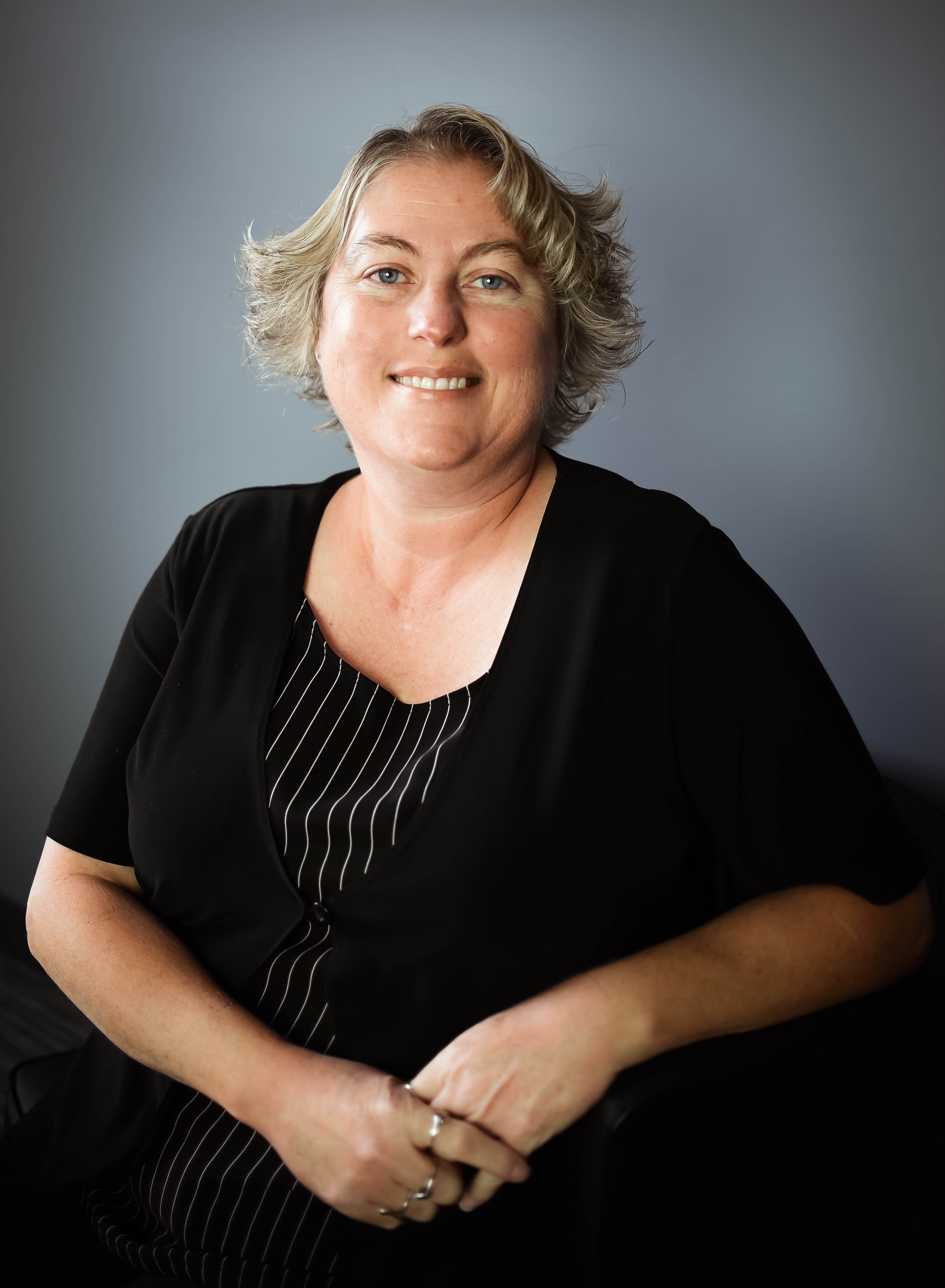 A woman with short, wavy blonde hair and blue eyes, smiling softly, wearing a black top with a striped inner piece, sitting against a plain gray background.
