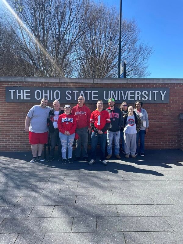 Group of nine people standing outside in front of a brick wall with a sign that reads 'The Ohio State University,' with bare trees and a clear blue sky in the background.