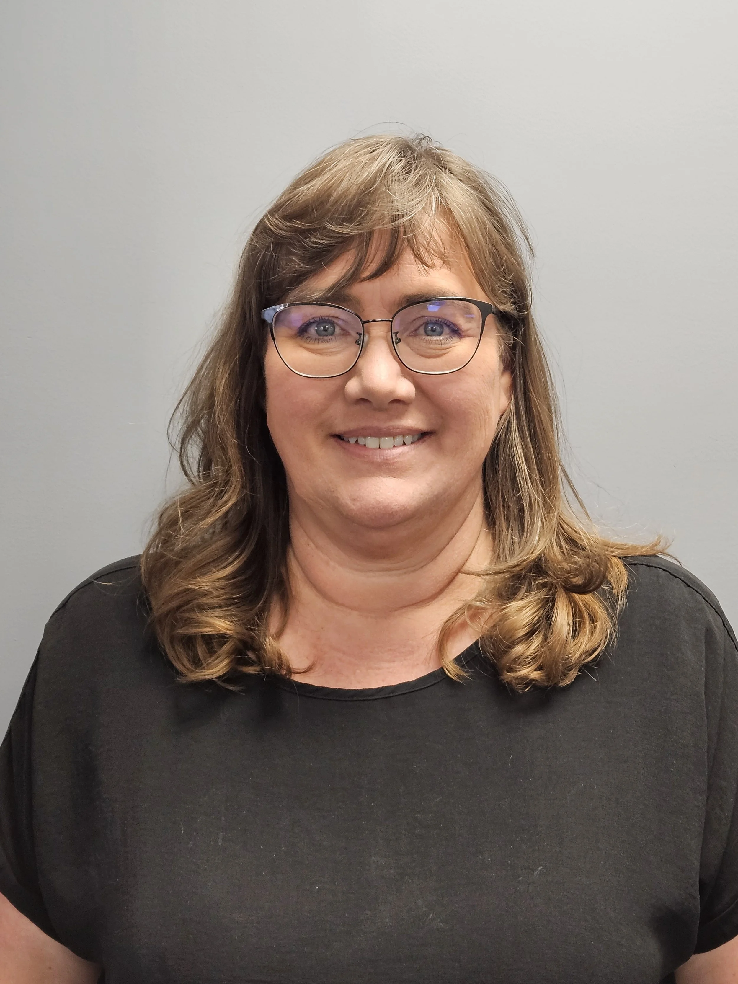 A woman with light brown, wavy hair and glasses, smiling and wearing a black top, standing against a plain light gray background.