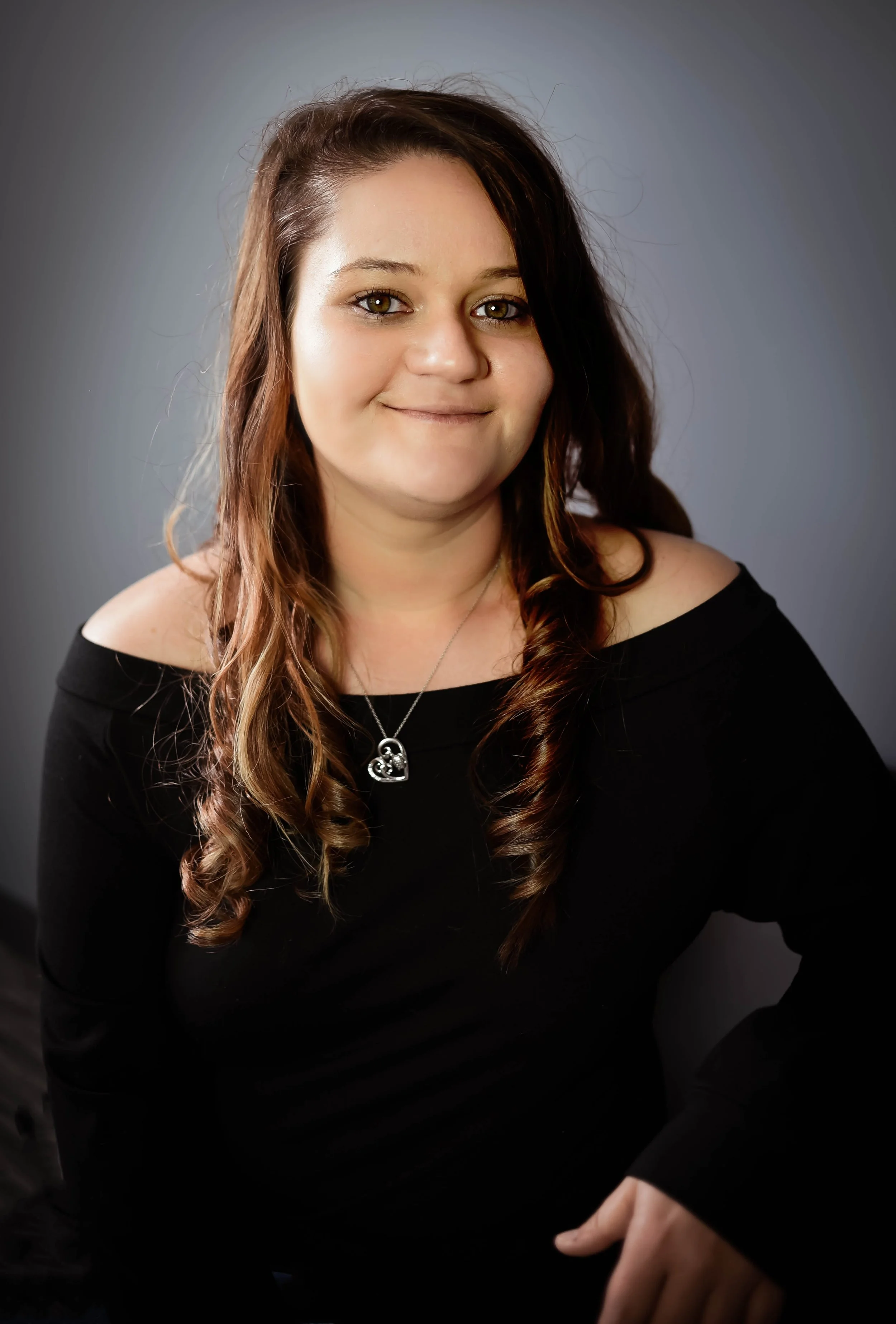 A young woman with long, wavy brown hair and hazel eyes, wearing a black off-the-shoulder top, smiling softly, with a gray background.