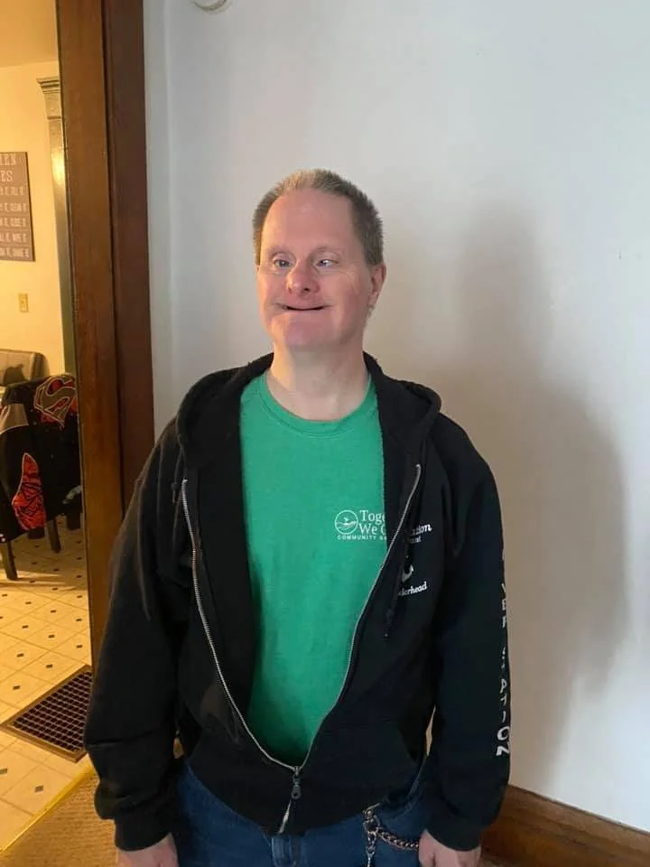 A man with short, light-colored hair and a wide smile, wearing a green T-shirt and a black zip-up jacket, standing indoors next to a white wall.