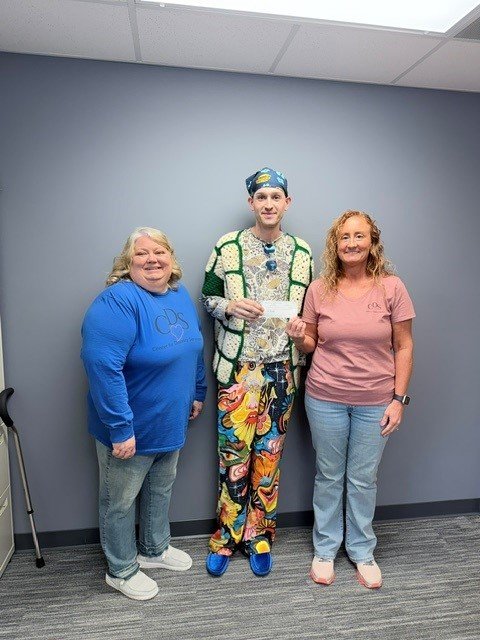 Three people standing indoors against a gray wall, smiling at the camera. The person in the middle is dressed in colorful, eccentric clothing, including a patterned jacket and pants, and a hat. The person on the left is wearing a blue sweatshirt and jeans, and the person on the right is wearing a pink shirt and jeans.