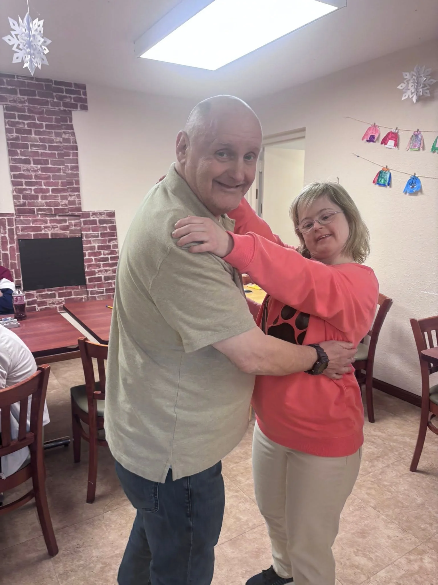 A man and a woman are dancing together in a social setting, smiling at each other. The man is bald and wearing a beige shirt and jeans. The woman has shoulder-length blonde hair, glasses, and is wearing a coral sweater and beige pants. The background