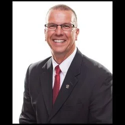 Portrait of a smiling man in a black suit, white shirt, and red tie, wearing glasses, against a white background.