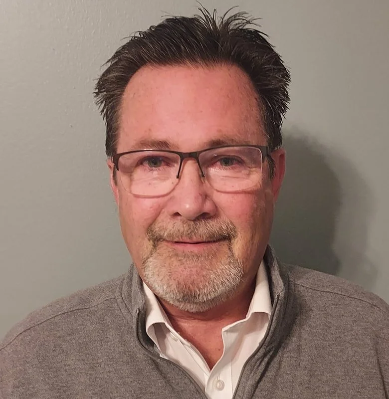 A middle-aged man with glasses, short dark hair, a beard, and a mustache, wearing a white collared shirt and a gray zip-up sweater, standing against a plain gray background.