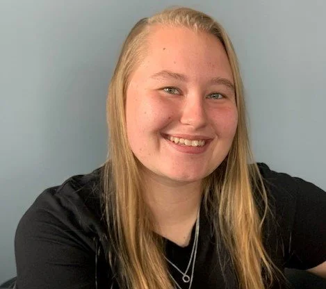 A young woman with long blonde hair smiling, wearing a black shirt, standing against a light blue wall.