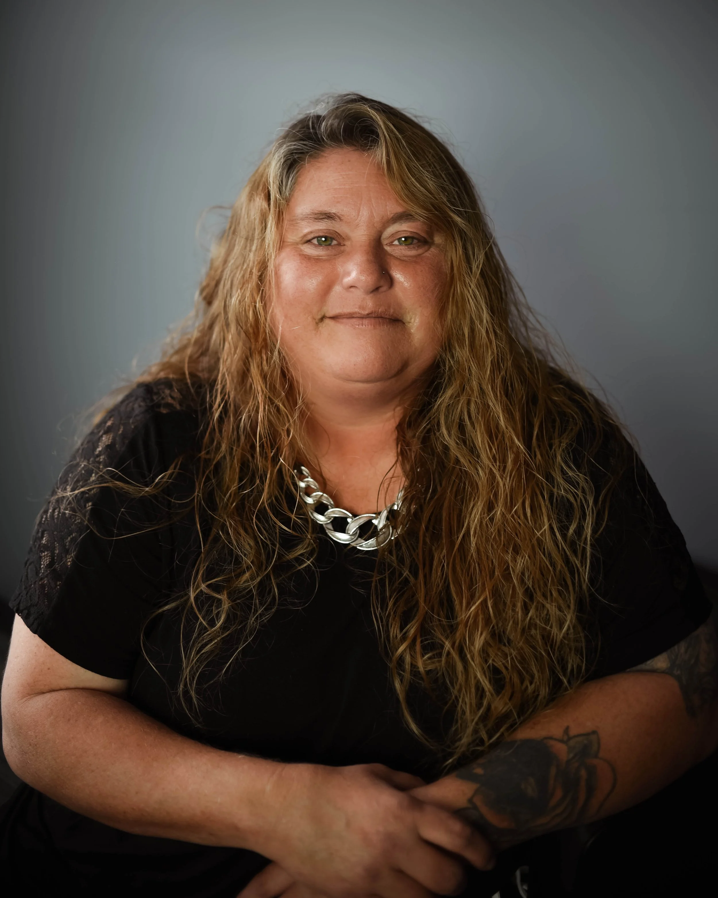A woman with long, wavy, blond hair and green eyes smiling, wearing a black shirt with a silver chain necklace, sitting with arms crossed against a plain background.