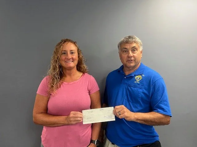 A woman and a man standing side by side against a gray wall. The woman is wearing a pink shirt and holding a check. The man is wearing a blue polo shirt with a logo on it.