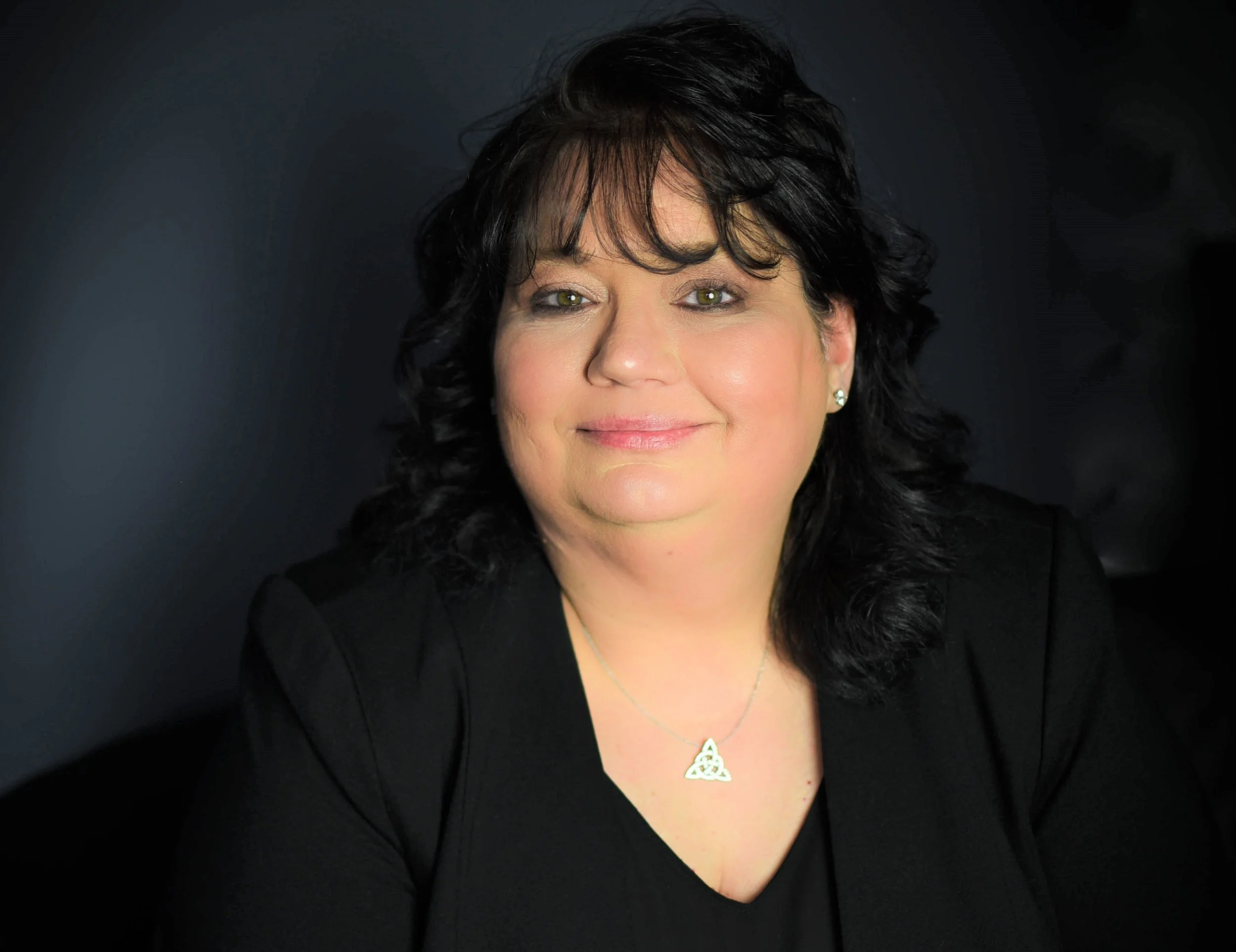 Close-up of a woman with short dark hair, green eyes, wearing a black top, a Celtic knot necklace, and earrings, against a black background.