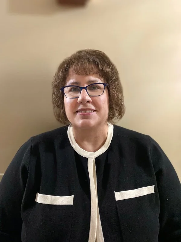 A woman with short, curly brown hair and glasses, wearing a black and white outfit, smiling and sitting against a plain beige wall.