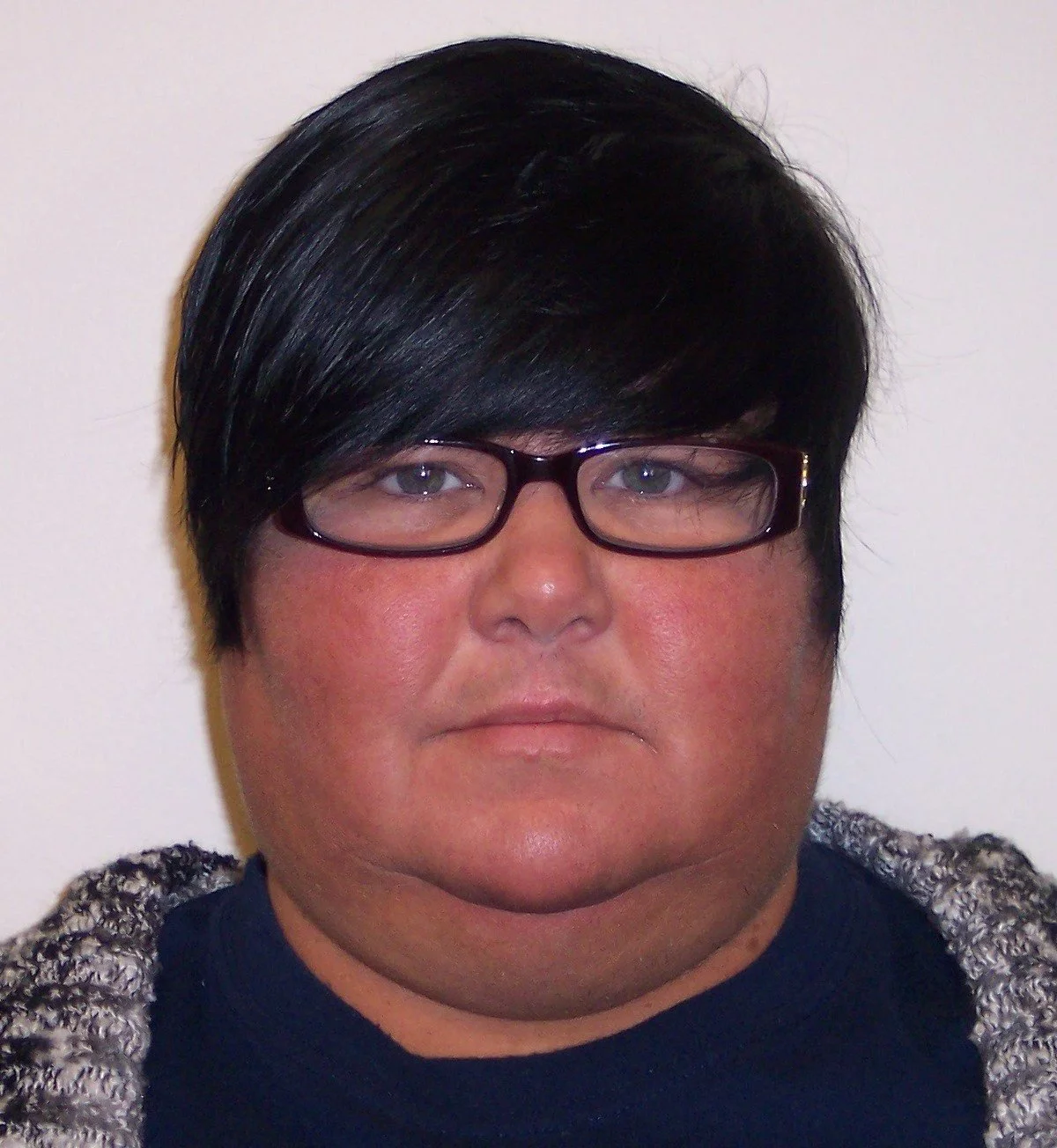 A person with short black hair, glasses, and wearing a dark shirt and a gray patterned sweater, looking directly at the camera against a plain light-colored background.