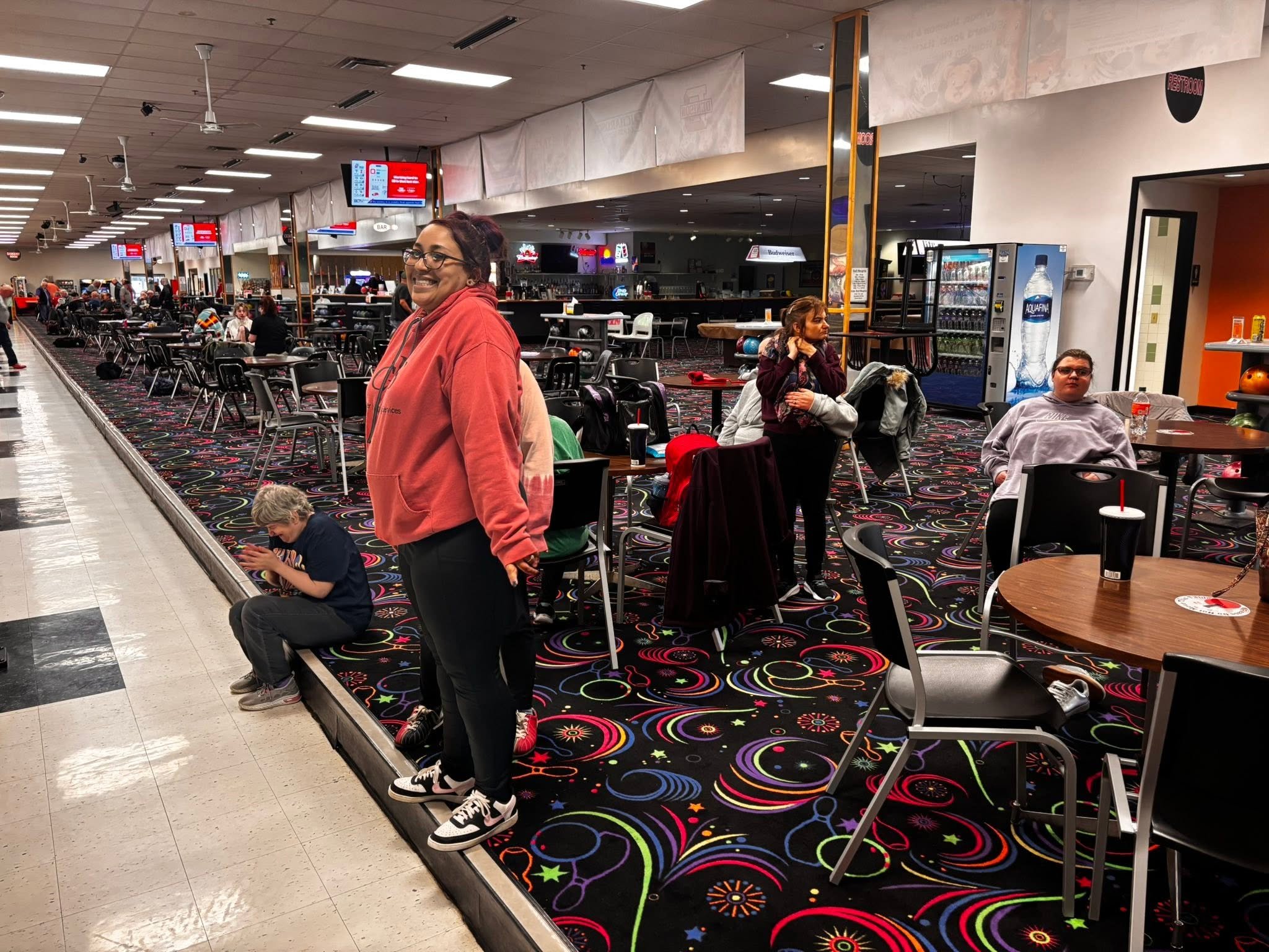 A large indoor space with colorful patterned carpet, several tables and chairs, and people sitting and standing. A woman with glasses and a red hoodie is smiling at the camera. There are vending machines, digital screens, and a bar area in the backgr