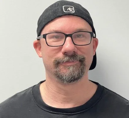 A man with glasses wearing a gray baseball cap and a black T-shirt, standing against a plain light-colored background.