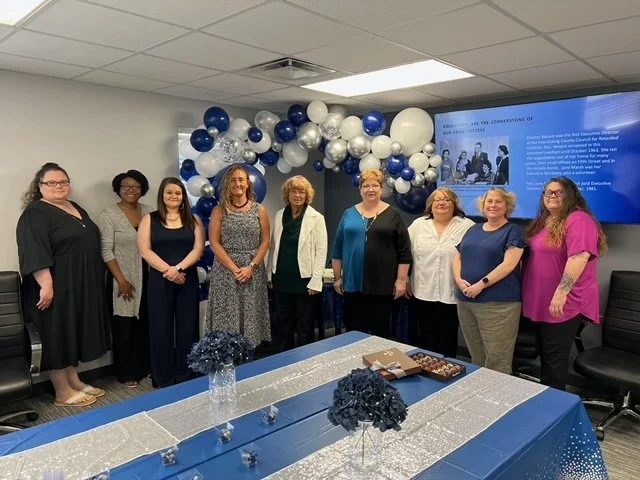 Group of women standing in a room decorated with blue and white balloons, with a table with blue and silver decorations in the foreground and a large screen displaying a presentation in the background.