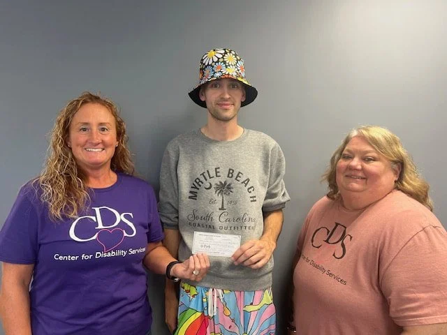 Three people standing together, two women and one young man in the middle, smiling for the photo. The woman on the left is wearing a purple shirt that says 'Center for Disability Services,' and the woman on the right is wearing a pink shirt with the same logo. The young man in the middle is wearing a colorful bucket hat, a gray Myrtle Beach sweatshirt, and colorful shorts, holding a piece of paper in his right hand.