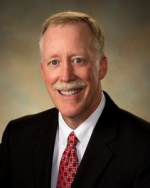 Portrait of a smiling middle-aged man with light hair, dressed in a black suit, white shirt, and red patterned tie, against a dark background.