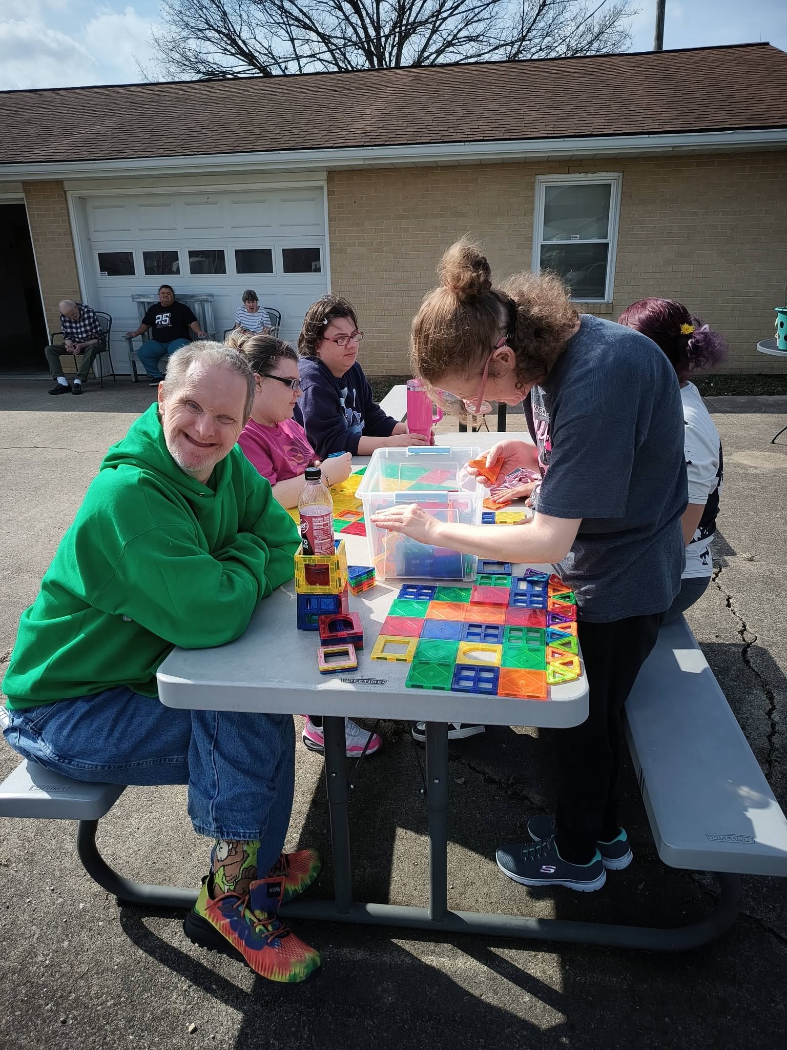 Group of people sitting at outdoor table playing with colorful geometric puzzles, with a house and garage in the background.