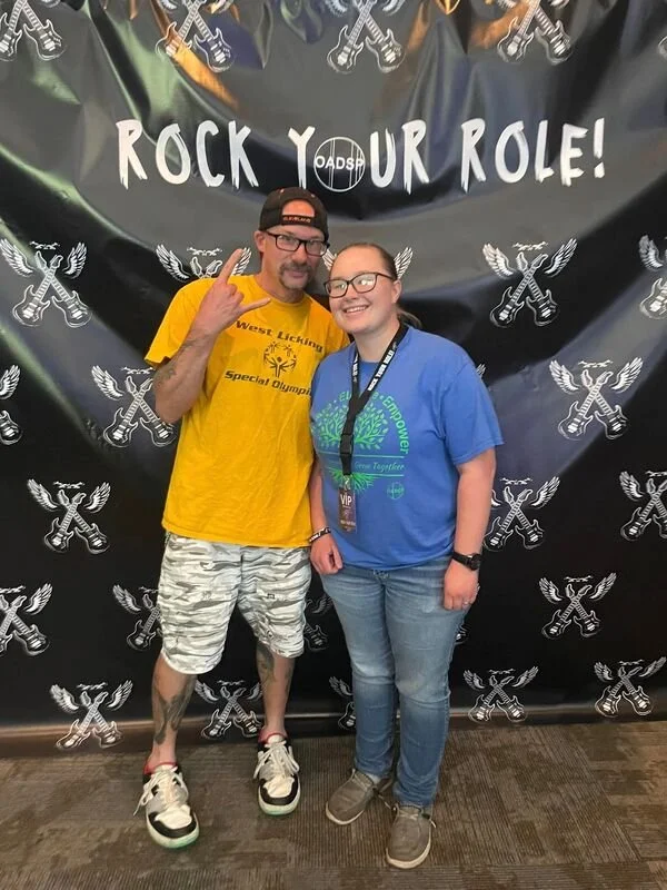 A man in a yellow shirt and a woman in a blue shirt standing together in front of a black backdrop with white logos and text that says 'ROCK YOUR ROLE!' The man is making a peace sign with his hand. The woman is wearing a lanyard and an event badge, 