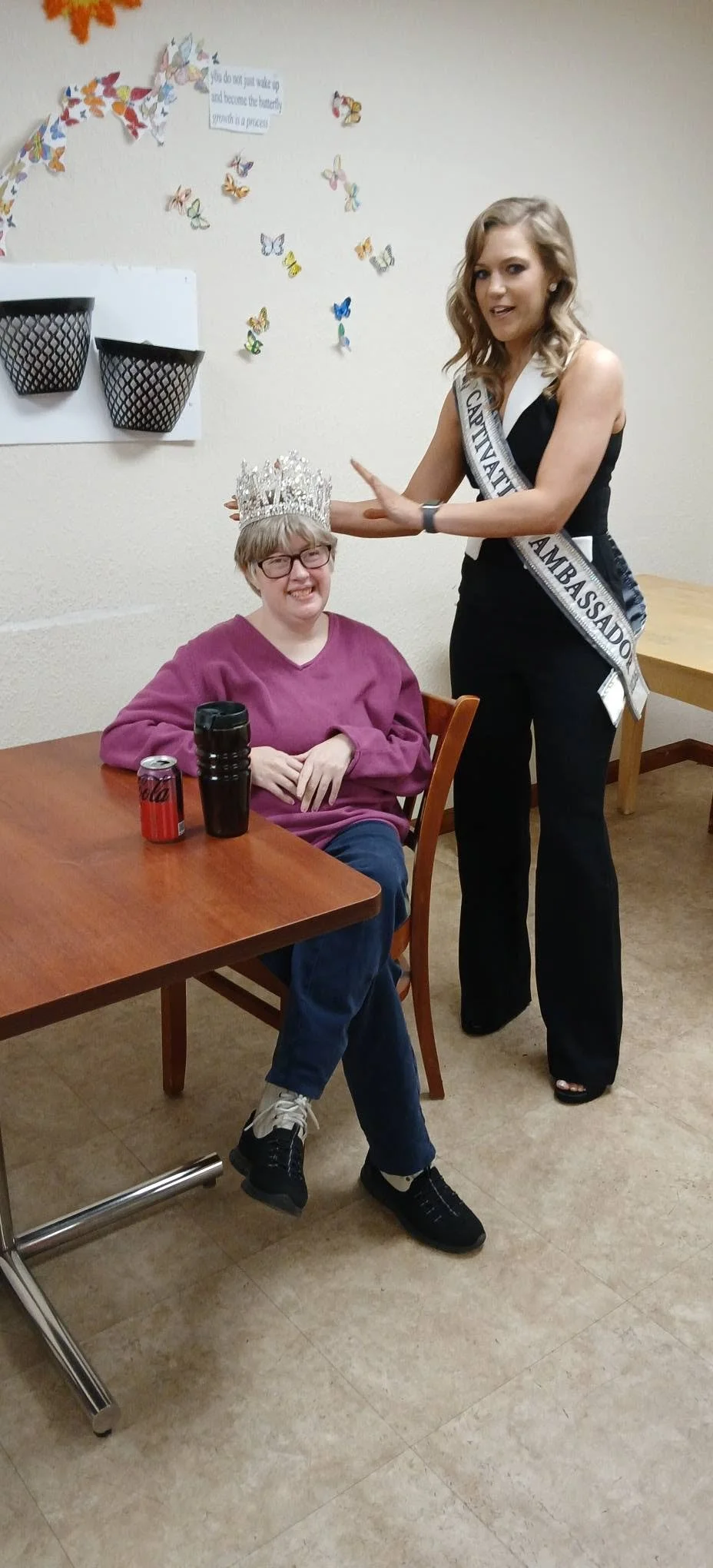 A woman wearing a sash and tiara is placing her hand on the head of a seated woman. The seated woman is smiling and wearing glasses, a purple sweatshirt, and jeans. There are butterflies and a quote on the wall behind them.