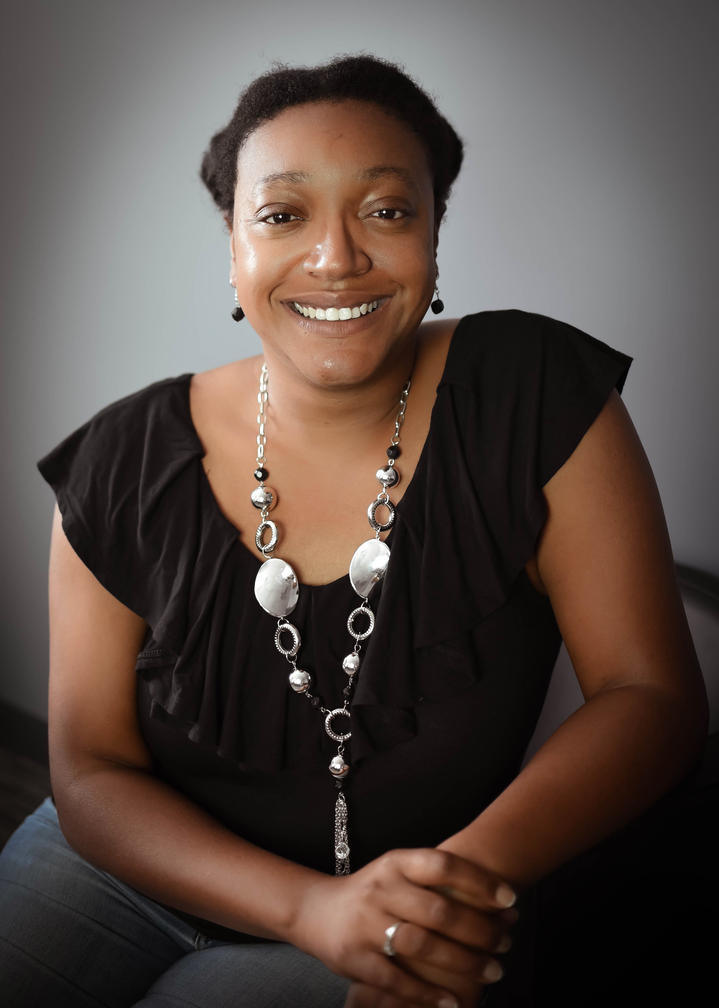A woman with dark skin, short dark hair, smiling, wearing a black top, silver and black jewelry, seated in front of a plain gray background.